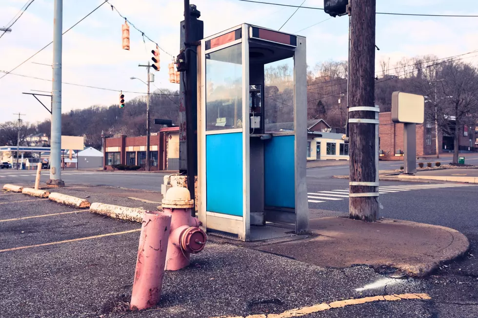 A classic vintage public phone booth with blue paneling standing on a street corner near a road intersection.