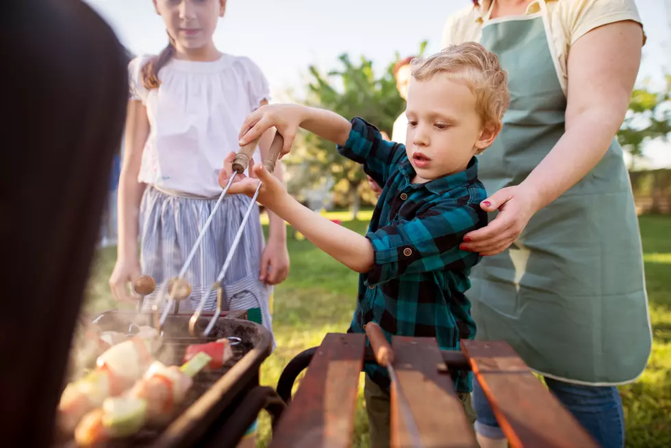 Focused little adorable toddler boy is carefully rotating meat and vegetables on a stick on a grill while being helped by his caring mother.