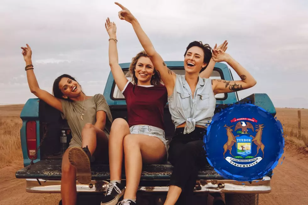 A group of three women sit on the tailgate of a moving pickup truck with the image of the Great Seal of Michigan seen in the lower right hand corner.