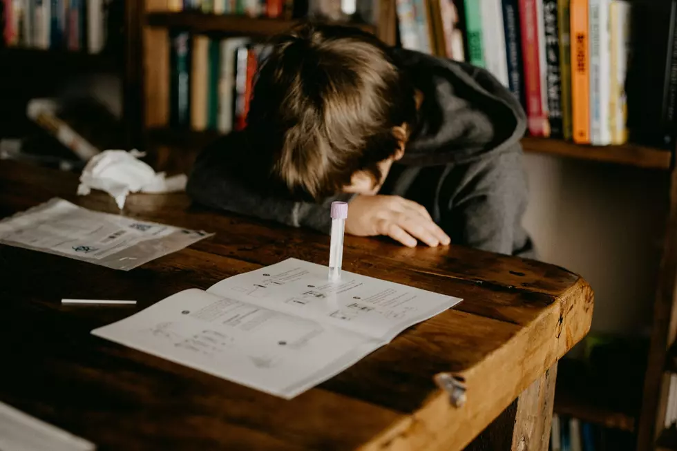 A child in a gray hoodie sits with his head down on a desk in school with a test tube holding a cotton swab in front of him.