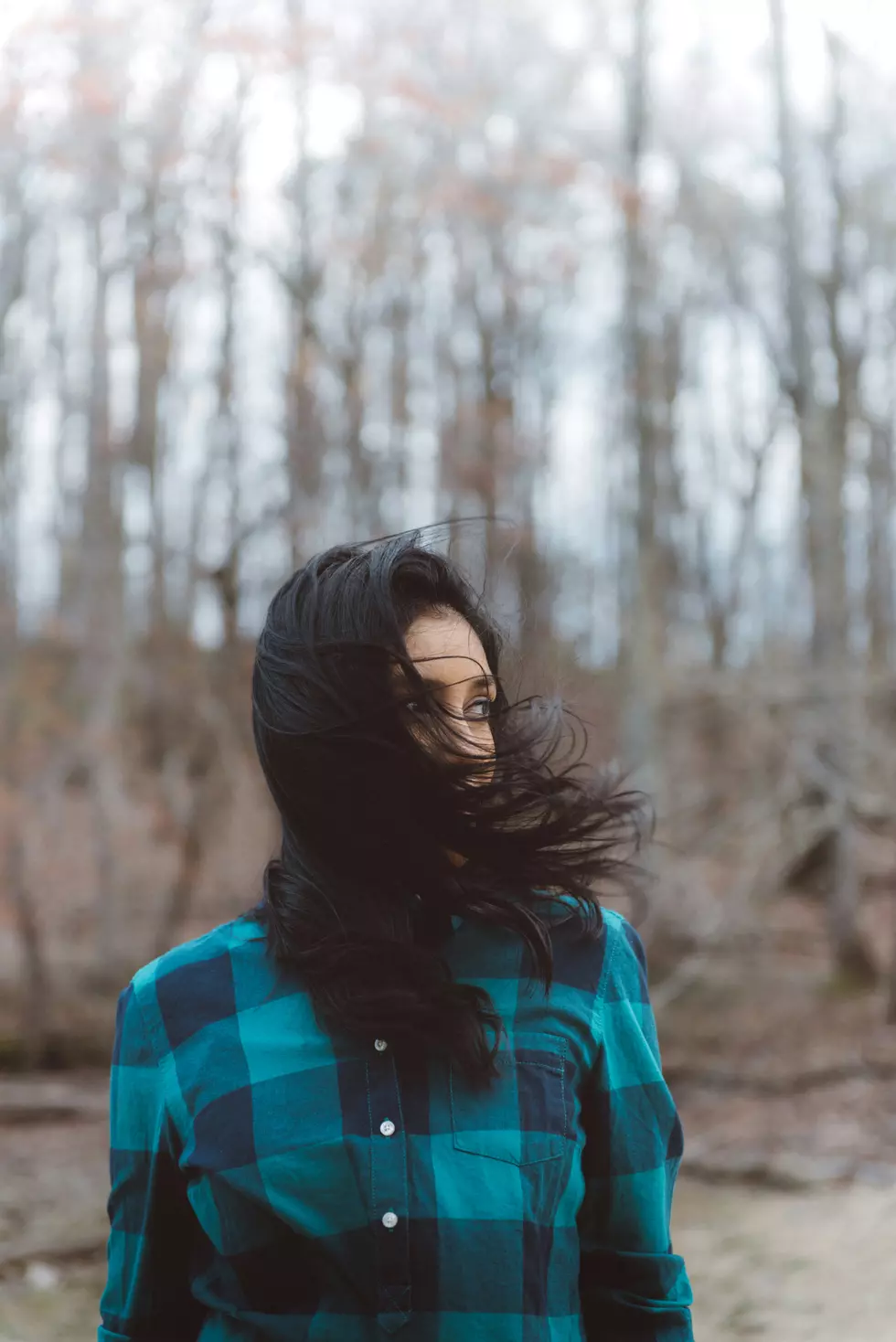 a woman in plaid with trees behind her and her hair blowing in the wind