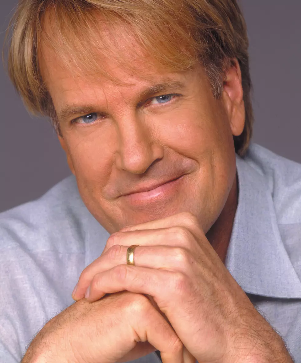 celebrity John Tesh wearing a blue shirt and smiling, hands folded.